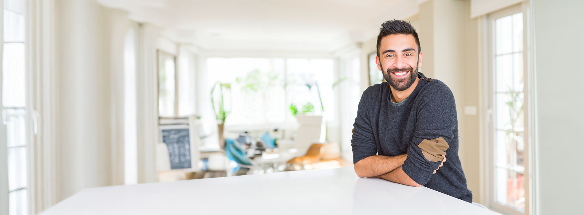 The image shows a man standing at a kitchen counter with a smile, wearing a dark top and light pants, against a blurred background of a kitchen interior.
