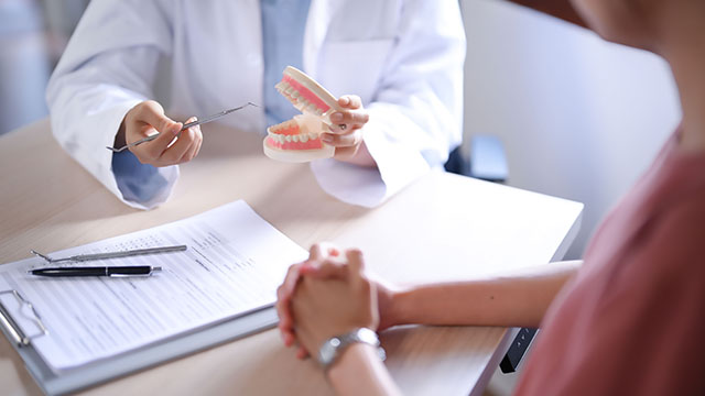 The image shows a dentist holding up a model tooth to a patient s mouth during an examination, with both individuals seated at a table with documents and a pen.